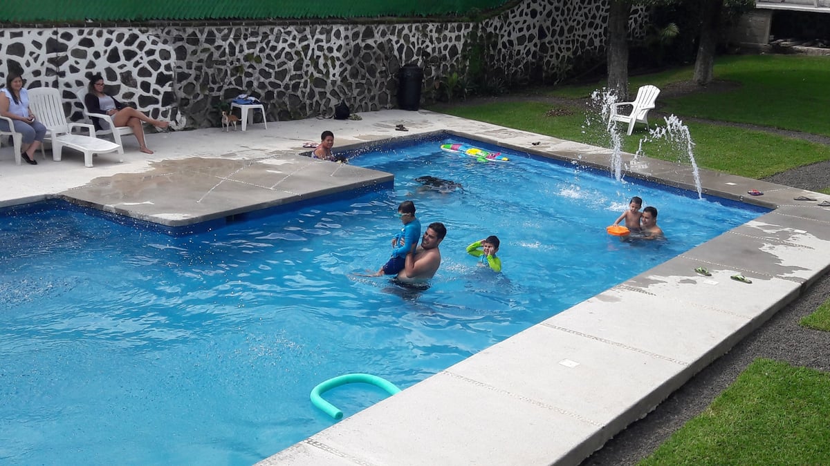 Personas nadando y relajándose en alberca rectangular con agua azul brillante, deck de concreto blanco y pasto verde
