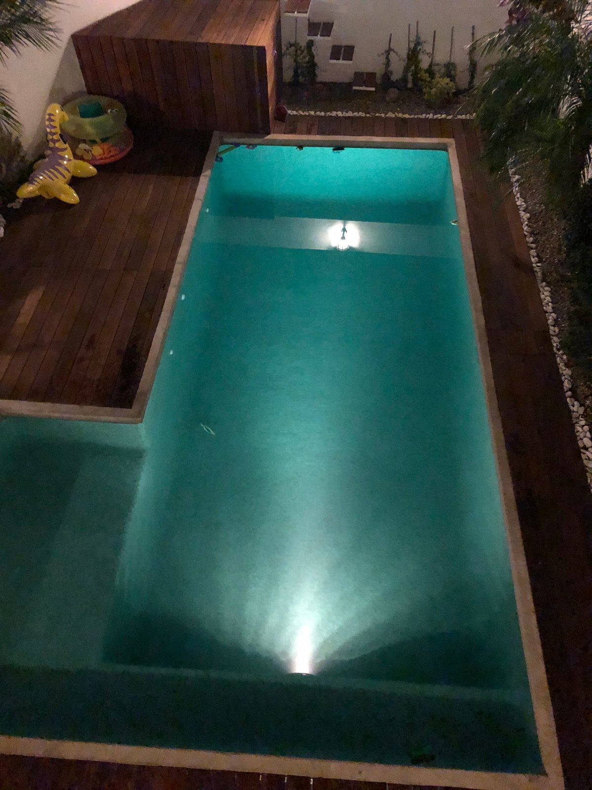 Vista aérea de alberca vacía turquesa con deck de ladrillo, camastros y plantas tropicales en jardín residencial