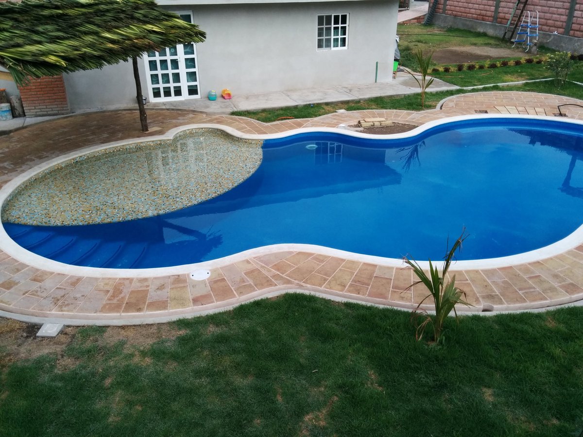 Alberca curva con área de chapoteo, deck de ladrillo y jardín con pasto junto a casa blanca