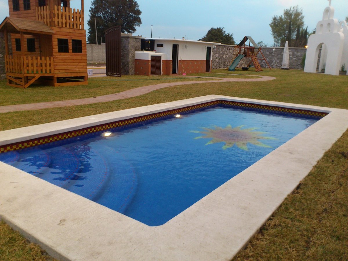 Alberca con agua azul y deck de concreto rodeada de pasto, casa de juegos de madera y construcciones