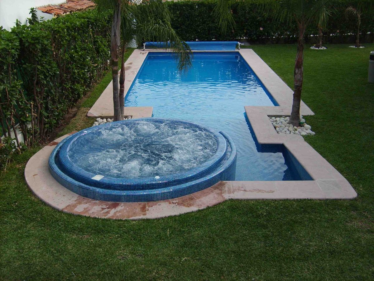 Jardín con alberca rectangular y jacuzzi circular en deck de concreto rodeado de pasto verde y setos