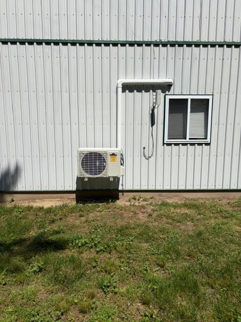HVAC outdoor unit mounted on metal building wall with window-mounted condenser unit and drainage pipe, surrounded by grass
