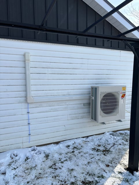 Outdoor HVAC unit mounted on white wall of covered structure with snowy ground below