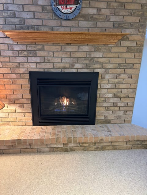 A brick fireplace with a black insert and wooden mantel, featuring a vintage neon clock sign above it