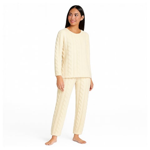 Woman wearing a cream-colored cable-knit pajama set with long sleeves and pants, standing barefoot against a white background