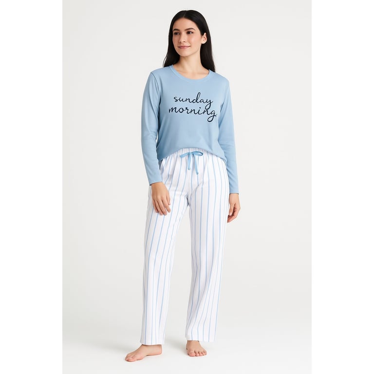 Woman wearing blue long-sleeve top with Sunday morning text and white striped pajama pants, standing barefoot against neutral background