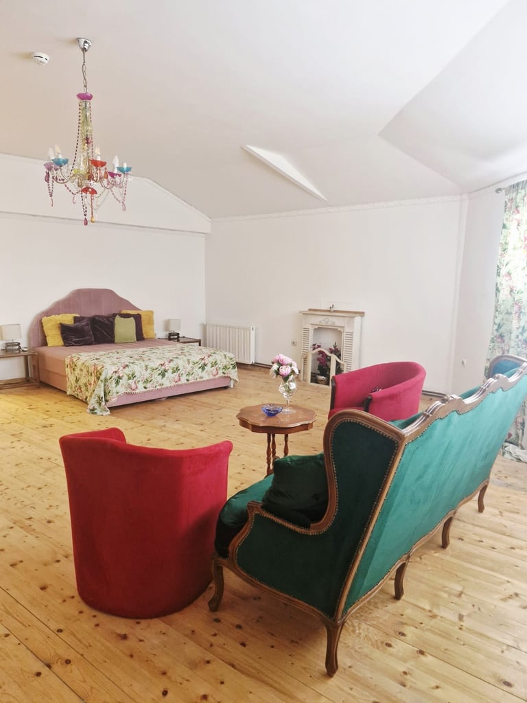 Colorful modern bedroom with wooden floor, upholstered chairs in red and turquoise, bed with pink headboard, ornate chandelier, and window with natural light