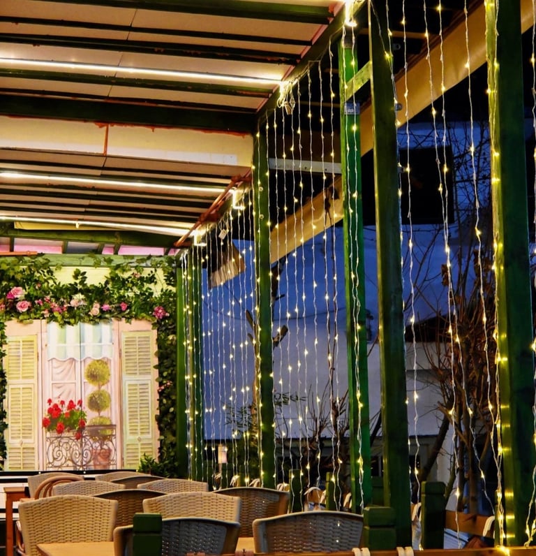 Modern restaurant interior with green metal frames, hanging string lights, floral decorations, and wicker chairs under linear ceiling lights