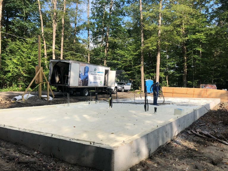Construction site with concrete foundation, mobile office trailer, and trees in background surrounded by wooden framing