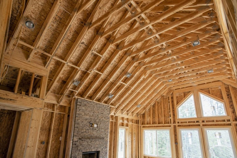 Interior of a new home under construction showing exposed wooden ceiling joists, framing, stone fireplace, and large windows
