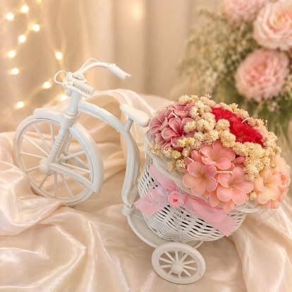 Decorative white tricycle with pink, red, and cream flowers in front basket on beige fabric with fairy lights