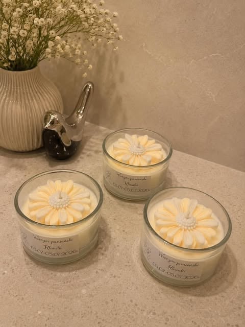 Three cream-colored candles with pale yellow daisy designs on a marble surface, with a ceramic vase of white flowers and decorative silver item nearby