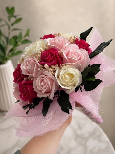 Hand holding a bouquet of red, pink, and cream roses wrapped in pink netting with black leaf accents