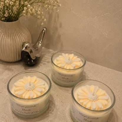 Three cream-colored scented candles with yellow daisy designs arranged on a surface next to a ribbed vase with white flowers
