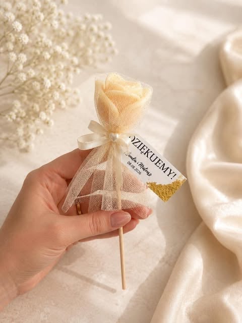 Hand holding a peach-colored decorative soap flower wrapped in tulle with a product label, displayed on white fabric backdrop with babys breath flowers
