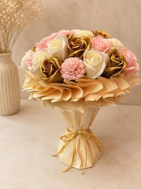 Elegant bouquet of golden, cream, and pink roses with carnations wrapped in gold ribbon, displayed in a glass vase