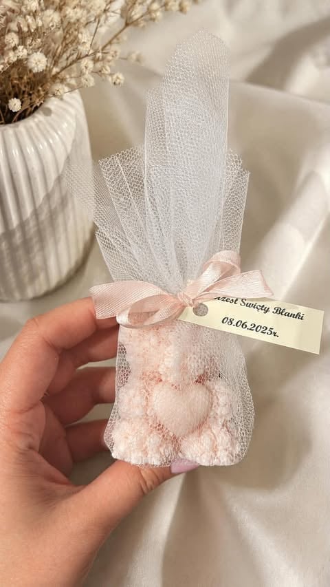 Hand holding small white mesh pouch with pink heart design and ribbon bow, labeled with white tag, next to dried flowers