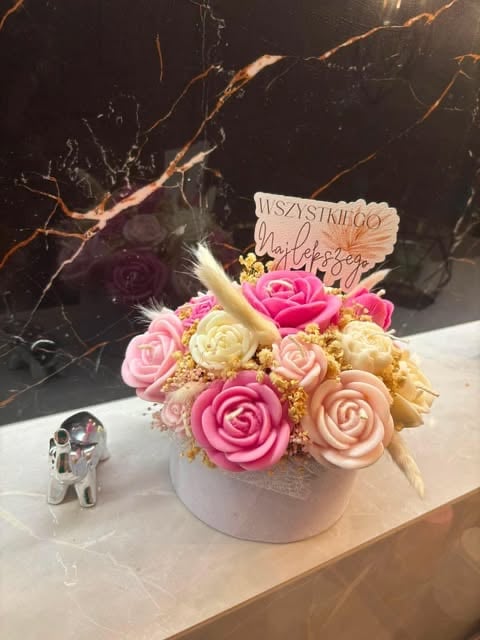 Decorative floral arrangement in white ceramic pot with pink and cream roses, gold accents, and a wooden sign on marble surface