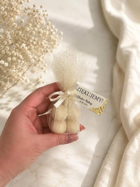 Hand holding a cream-colored tulle pouch with thank you tag, surrounded by babys breath flowers and white fabric