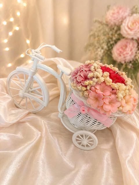 Decorative white tricycle with basket filled with pink, coral, and red flowers on champagne fabric with fairy lights and blooms in background