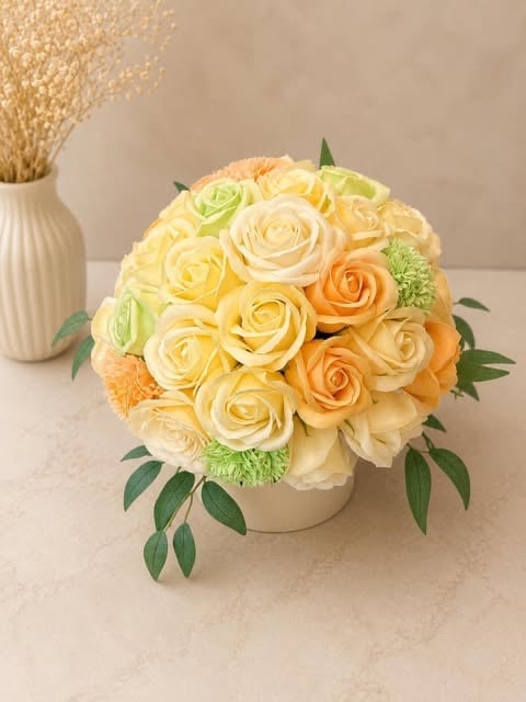 Arrangement of yellow, cream, and orange roses with green carnations in a white ceramic vase on a beige surface