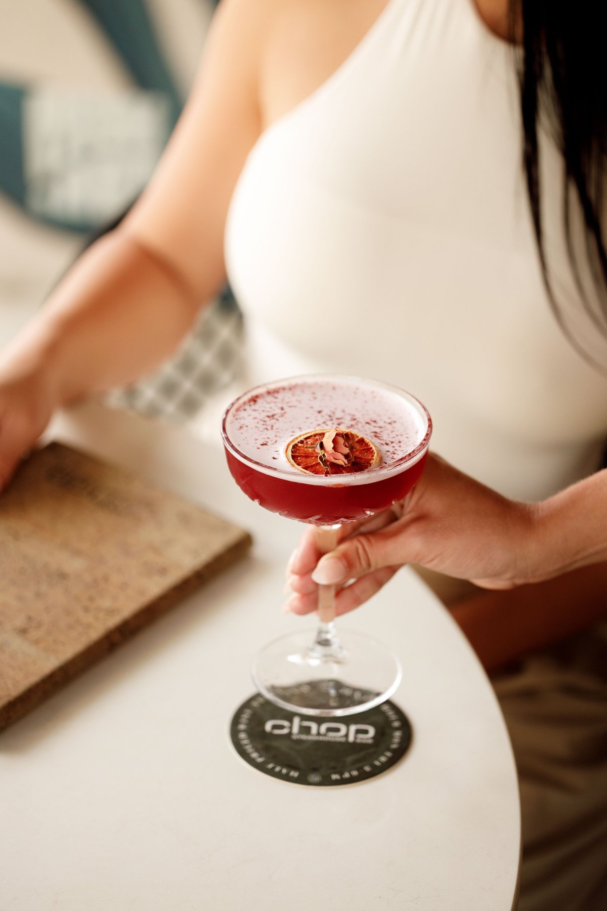 Woman in white clothing holding a red cocktail with foam top and grapefruit garnish at a bar