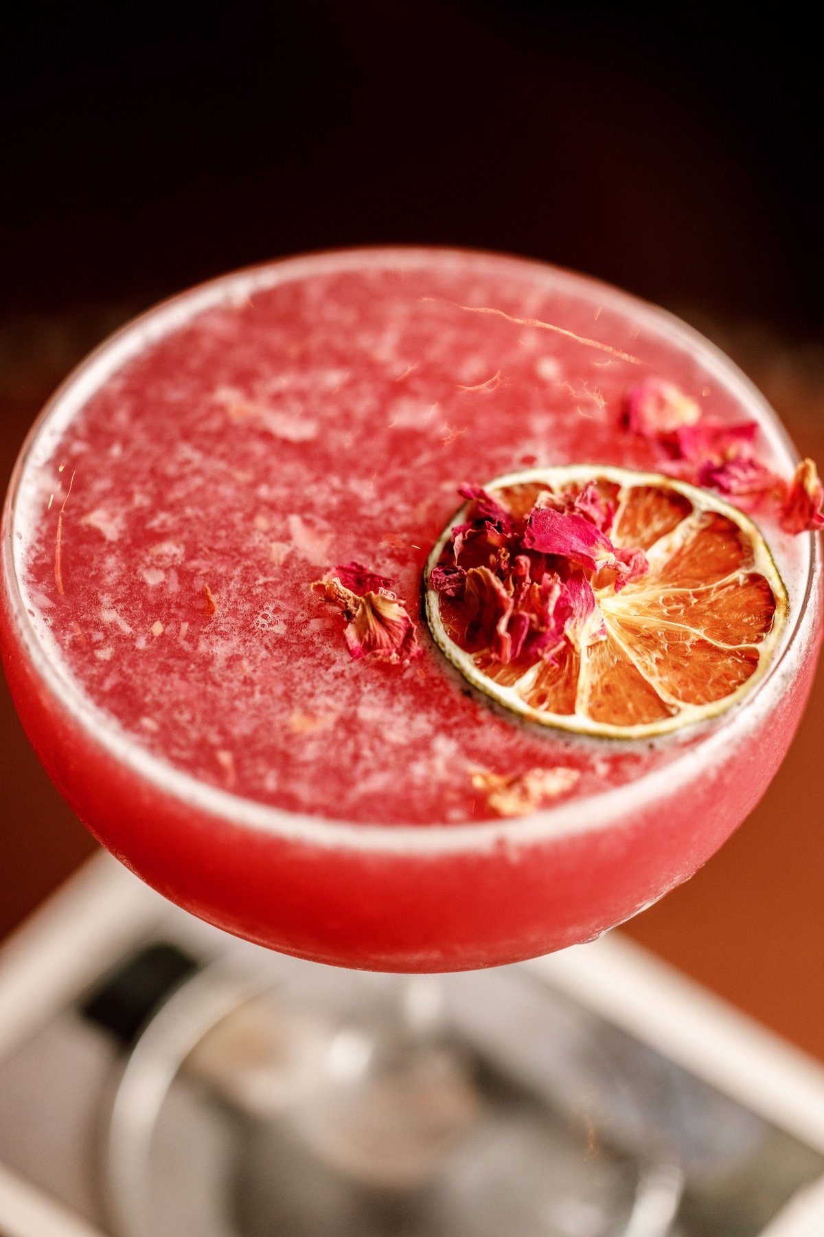 Pink cocktail with crushed ice, dried citrus slice and rose petals garnish on blurred background
