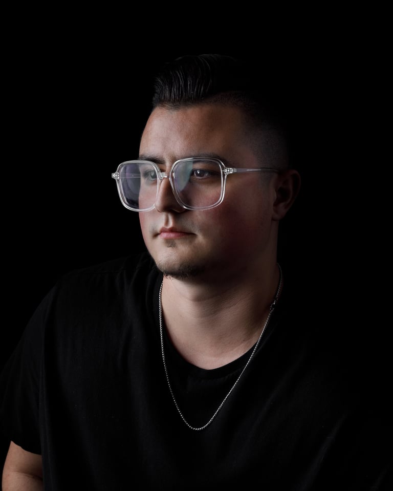 Man wearing clear-framed glasses and a black t-shirt with chain necklace against a dark background