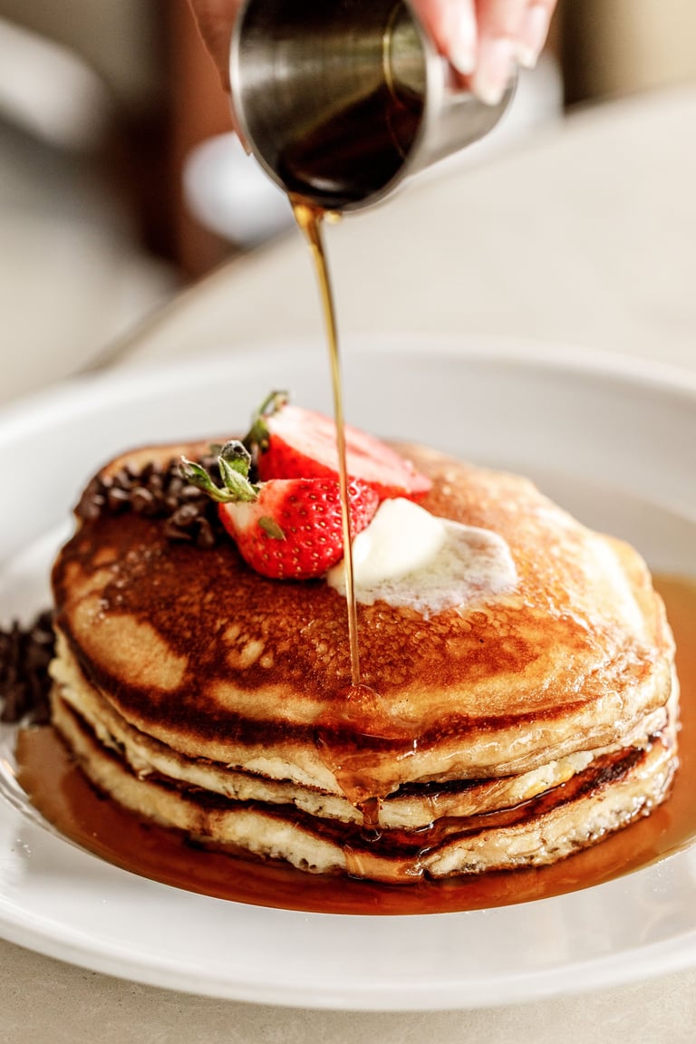 Maple syrup being drizzled over a stack of golden pancakes topped with berries and whipped cream on a white plate