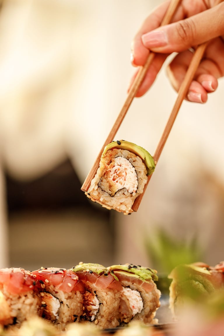 Hand holding chopsticks with a piece of sushi roll containing rice and ingredients above a plate of sushi