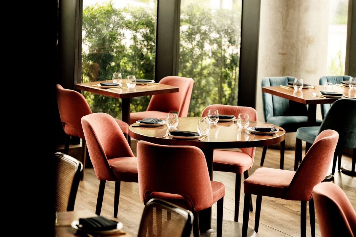 Modern restaurant interior with round black tables, rust-colored upholstered chairs, place settings, and large windows overlooking greenery
