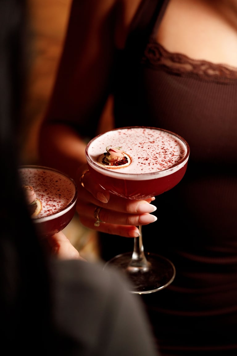 Person holding two cocktails with cocoa powder garnish and foam in warm golden lighting