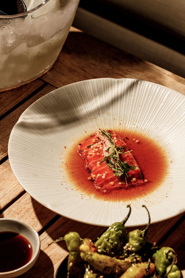 Sliced raw tuna arranged in a circle on a white plate with red sauce, garnished with microgreens and sesame seeds, served with a drink and peppers on a wooden table.