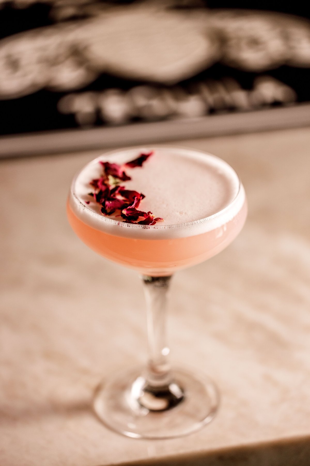 Elegant cocktail with coral liquid, white foam top, and dried rose petals garnish on a stem glass