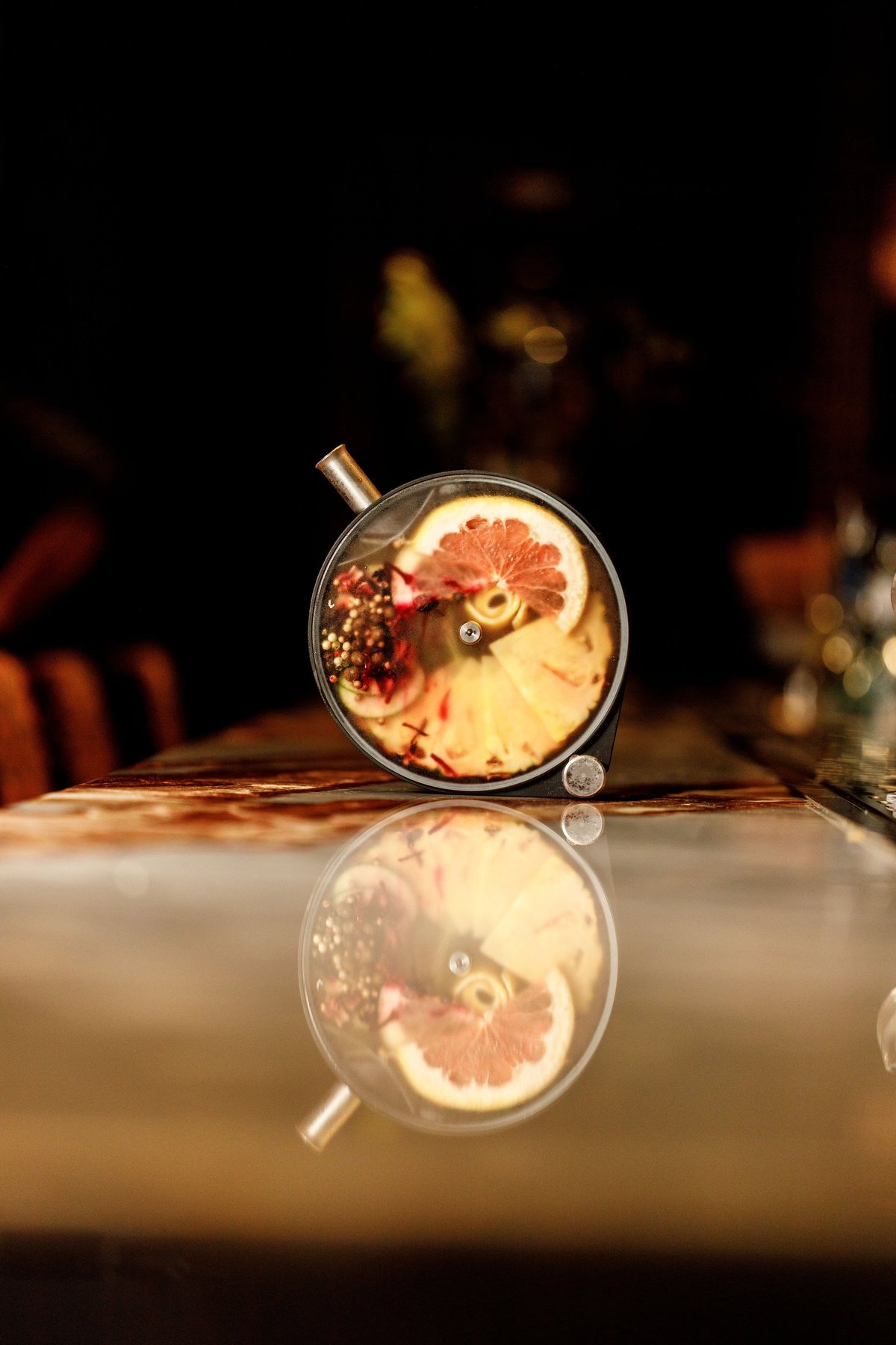 Overhead view of a clear glass mug containing a cocktail with grapefruit slice and pomegranate seeds, reflecting on a polished surface against a dark background