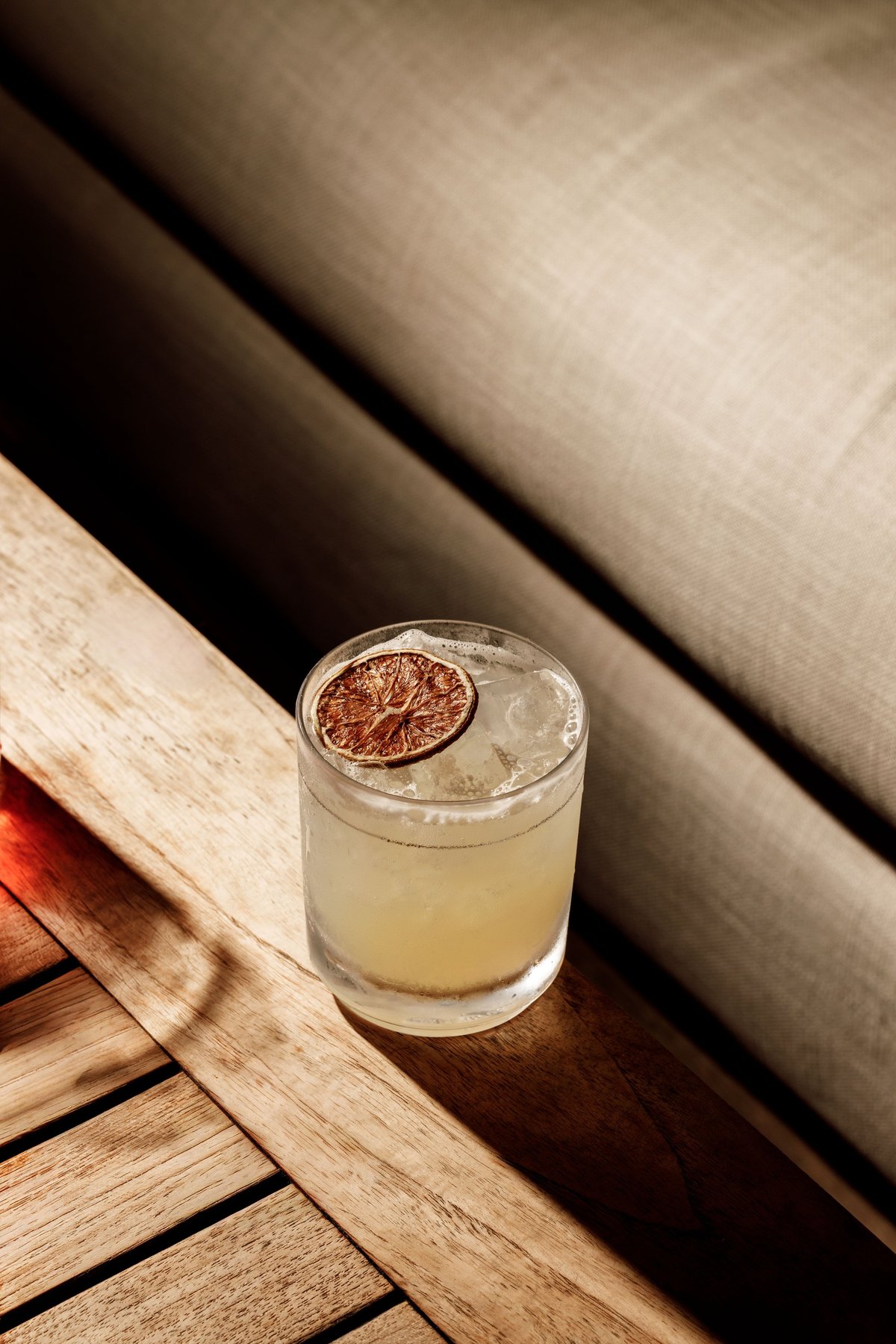 Glass of pale yellow cocktail with ice and dried citrus slice garnish on wooden surface