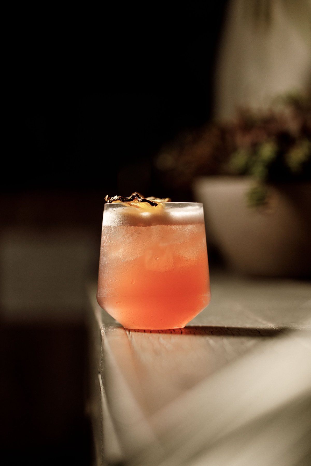 A coral-colored cocktail with ice and a gold leaf garnish sits on a marble surface in a dimly lit setting