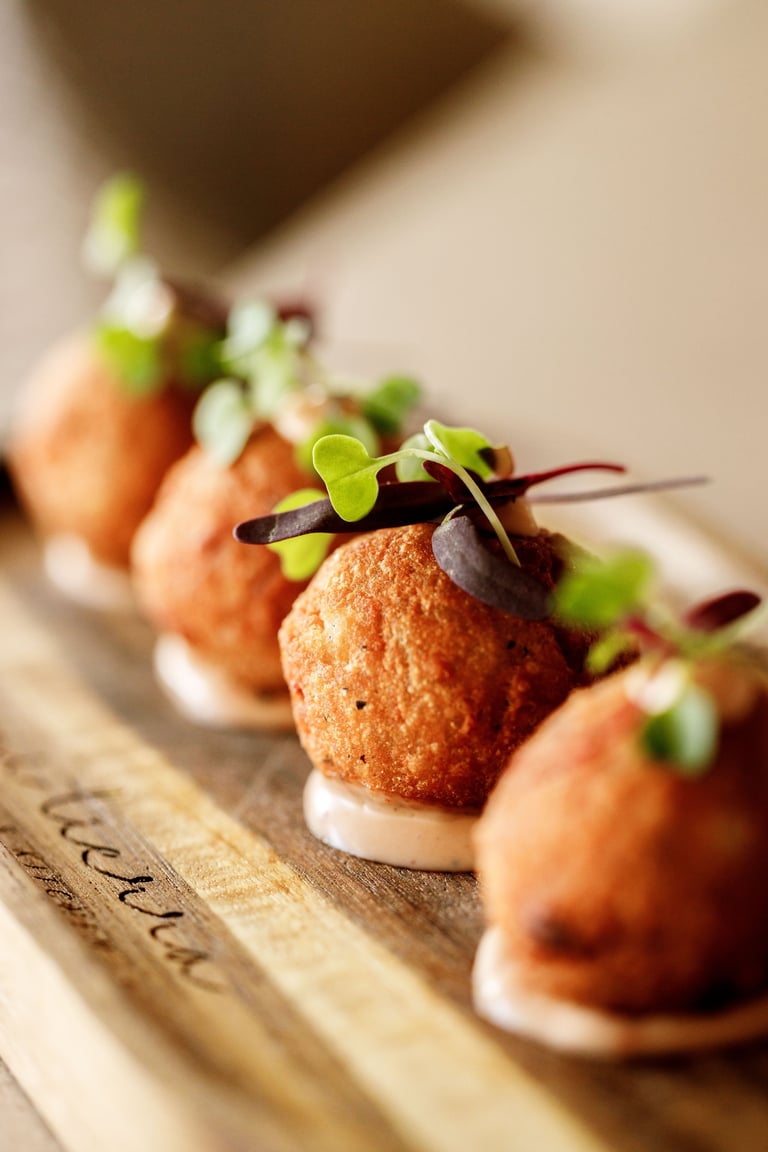 Row of gourmet appetizers with golden-brown fried spheres, white cream base, and fresh green microgreens and dark red garnish on wooden board