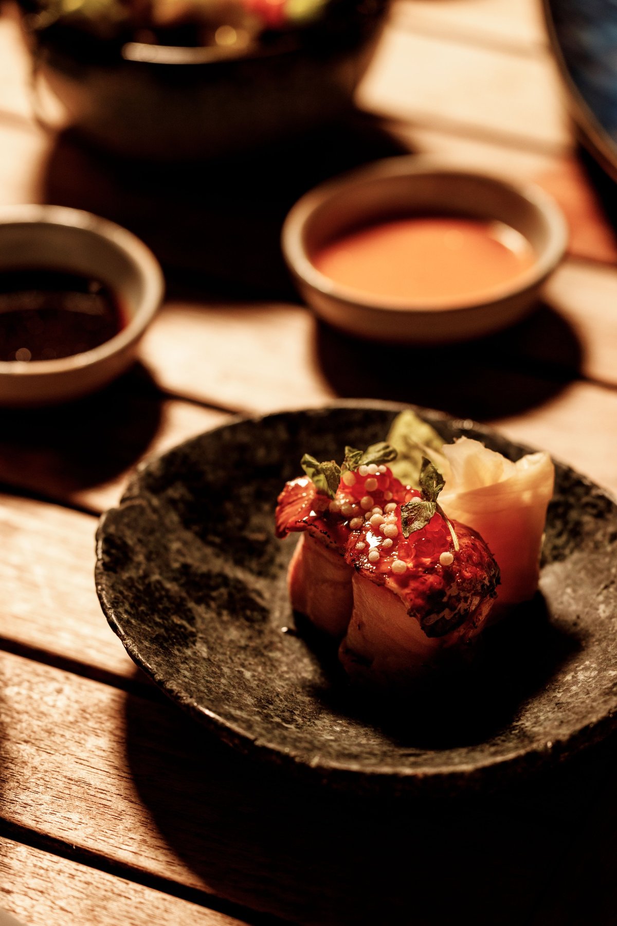A plated appetizer with red tuna, white garnish, and microgreens on a dark ceramic dish, with sauce bowls in the background.