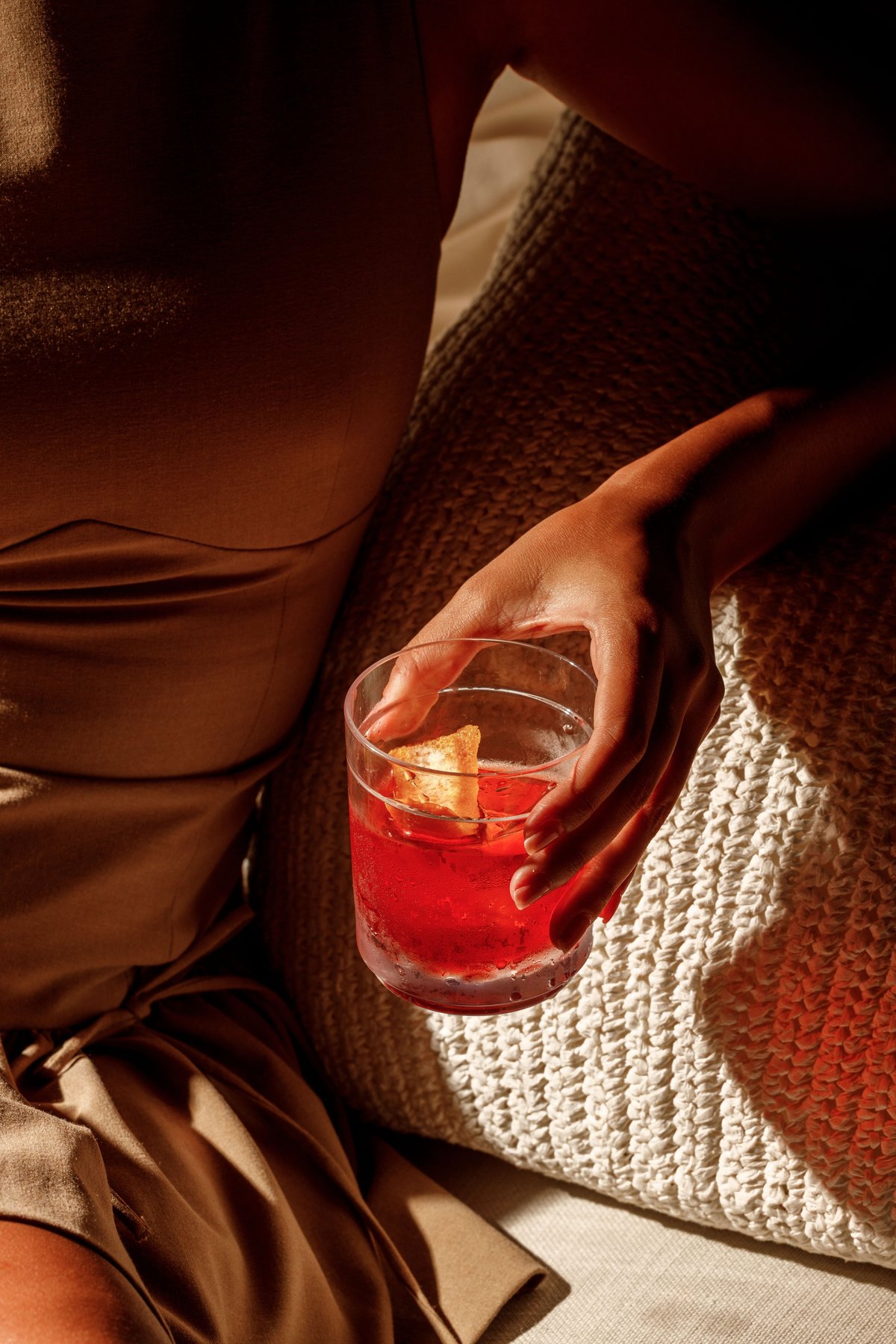 Person in cozy sweater holding glass of red cocktail with ice against warm-toned background