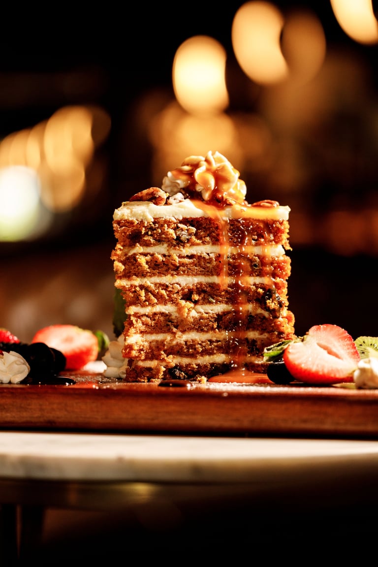 Layered chocolate cake slice topped with caramel and nuts on a wooden board with blurred warm lights in background