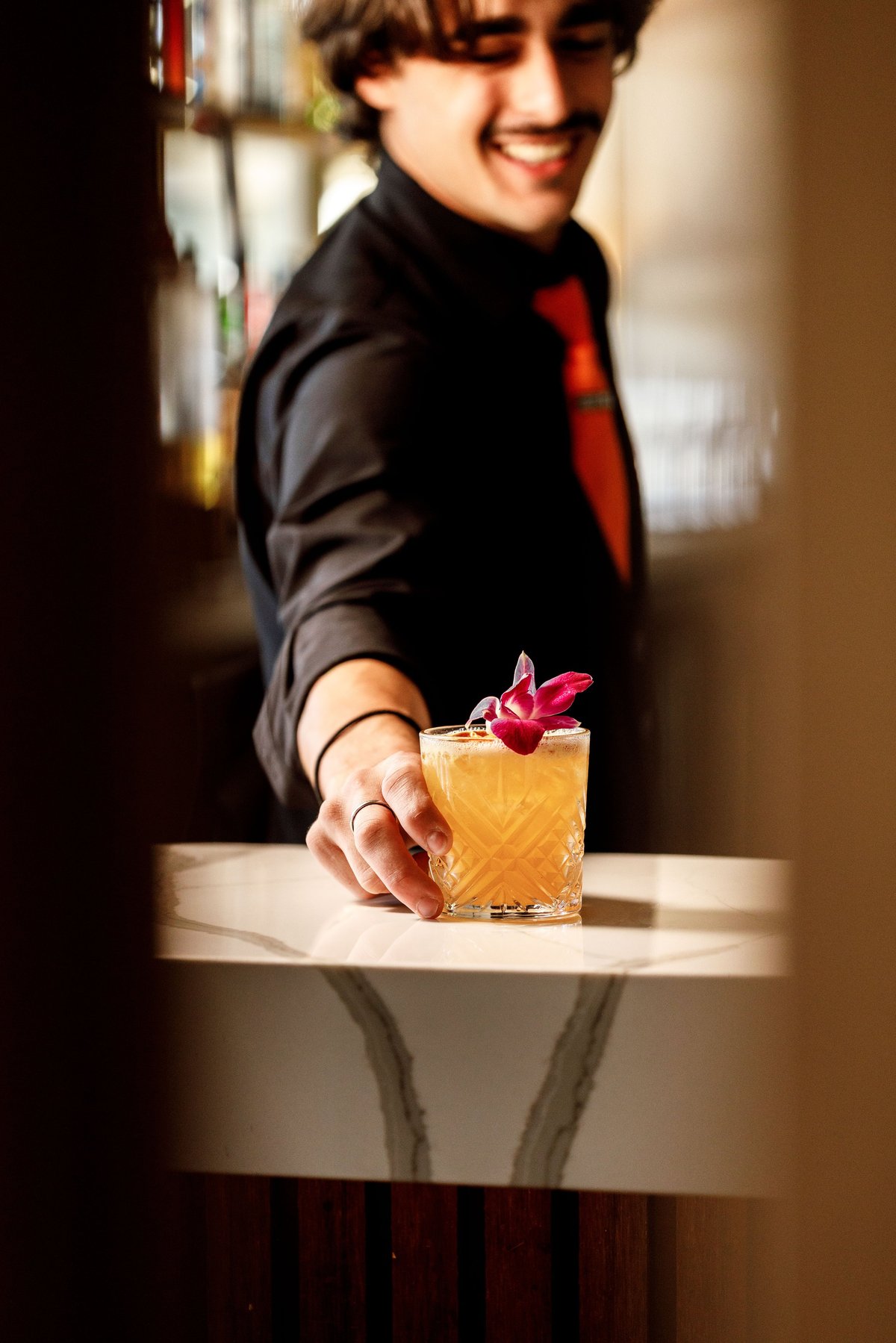 Bartender in black jacket and red shirt smiling while serving a golden cocktail garnished with a pink orchid flower