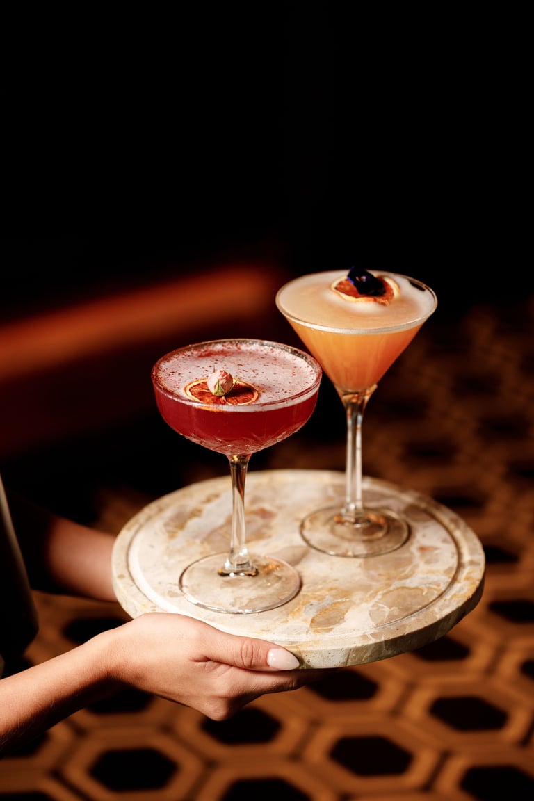 A hand holds a marble coaster with two cocktails: a red drink with a cherry and an amber-colored cocktail with a dark garnish.