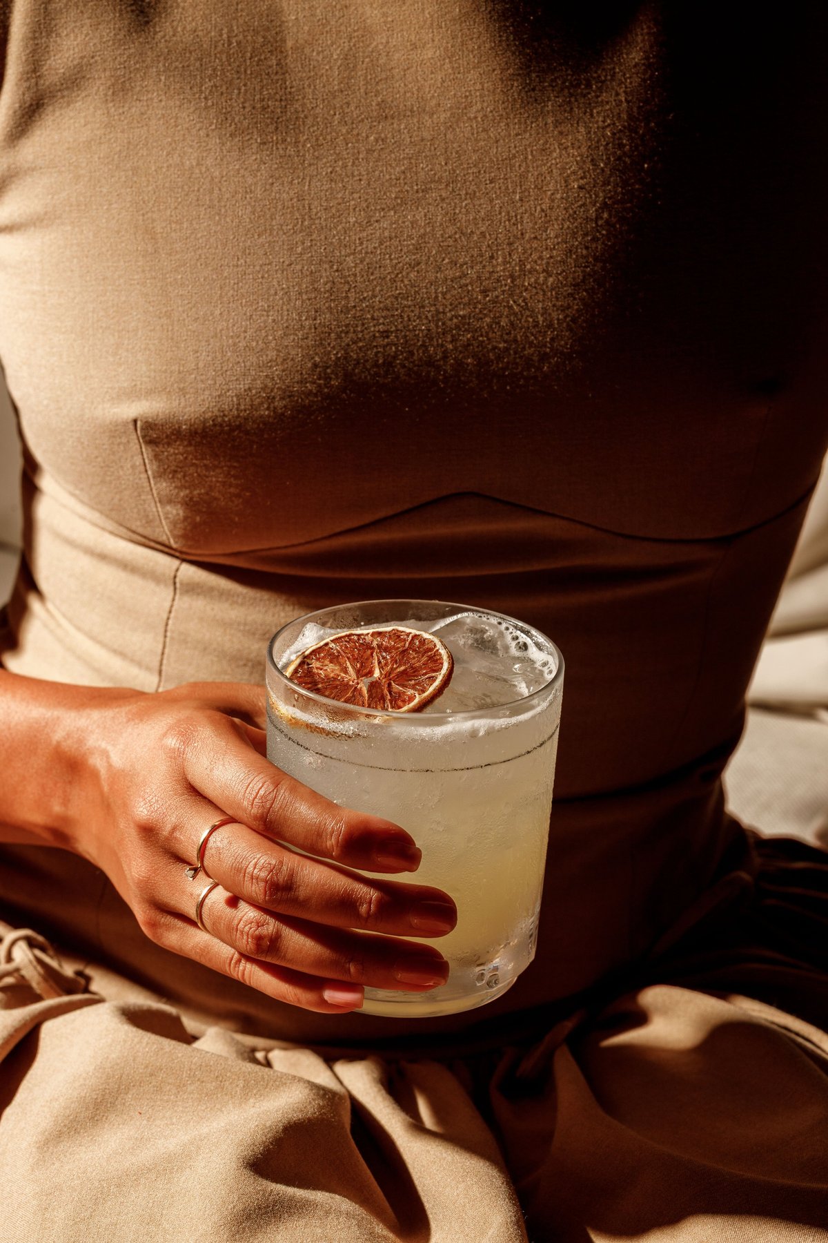 Person in beige clothing holding a glass of clear cocktail with ice and a grapefruit slice garnish