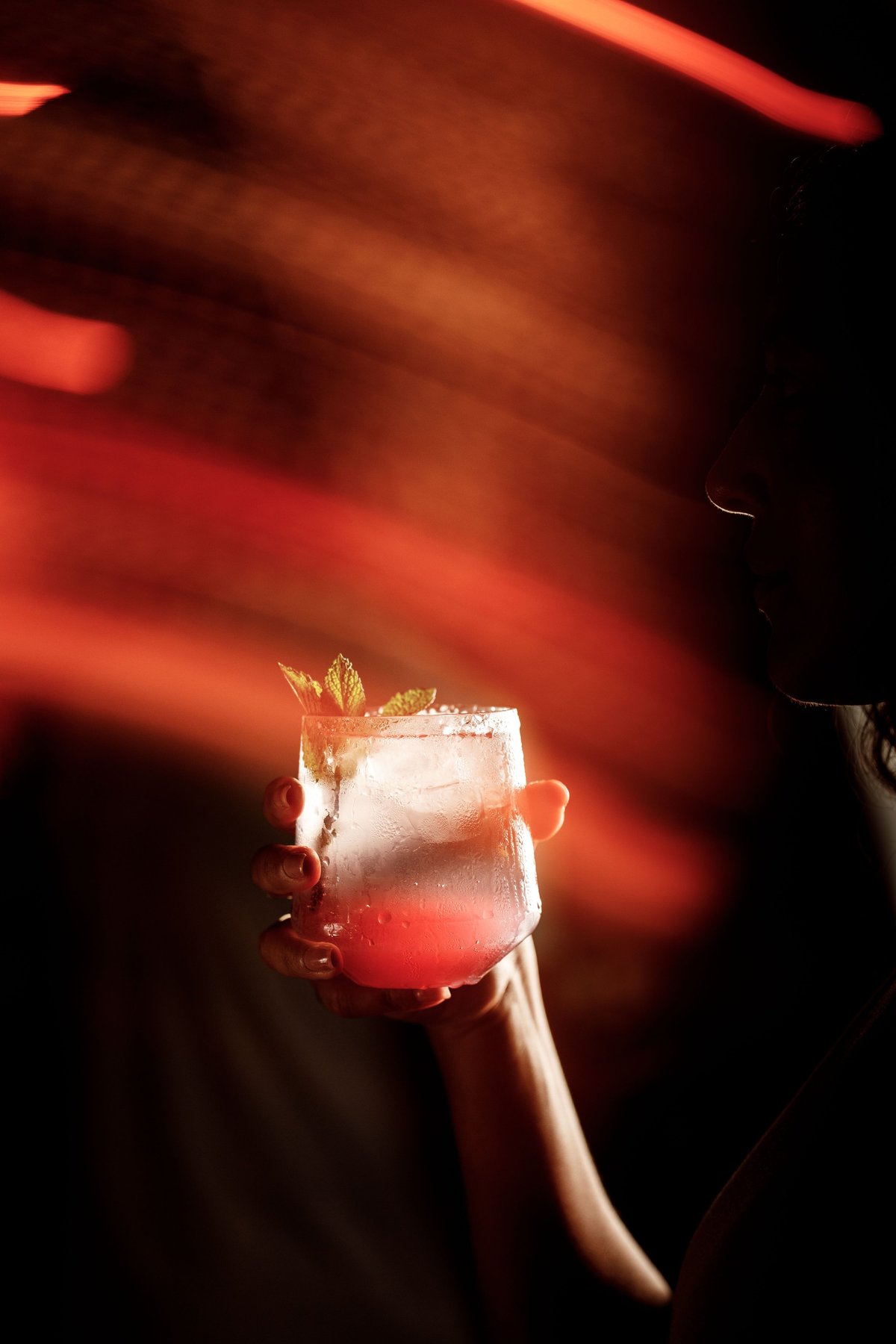 Hand holding a cocktail with ice and mint garnish against a warm red and orange glowing background