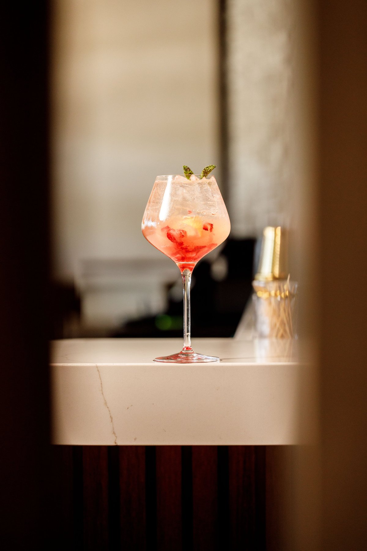 A coral-colored cocktail with ice and fresh herbs garnish in a wine glass on a marble counter