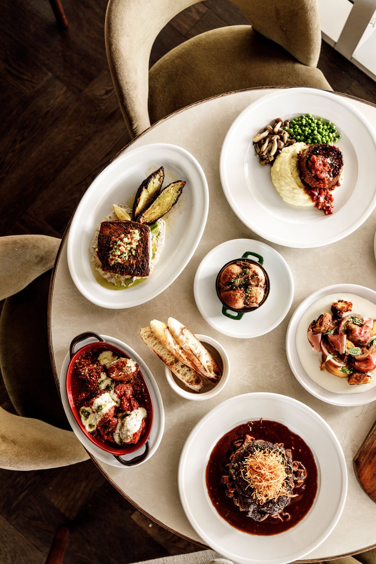 Overhead view of plated fine dining dishes on a round table with tan upholstered chairs, featuring prepared meats, vegetables, and garnishes on white plates.