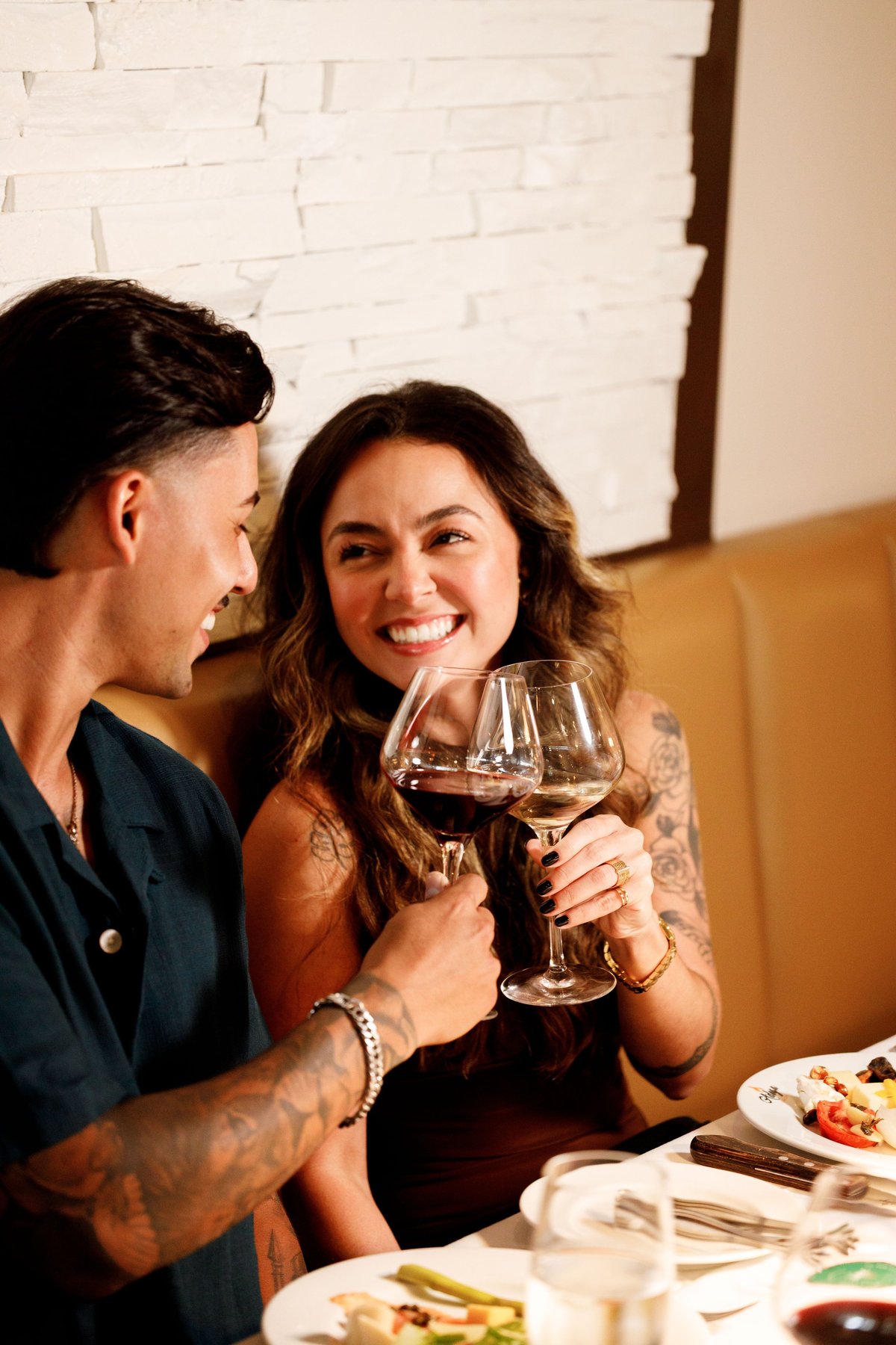 A man and woman toasting with wine glasses at a dinner table, smiling at each other in a warm, intimate moment.