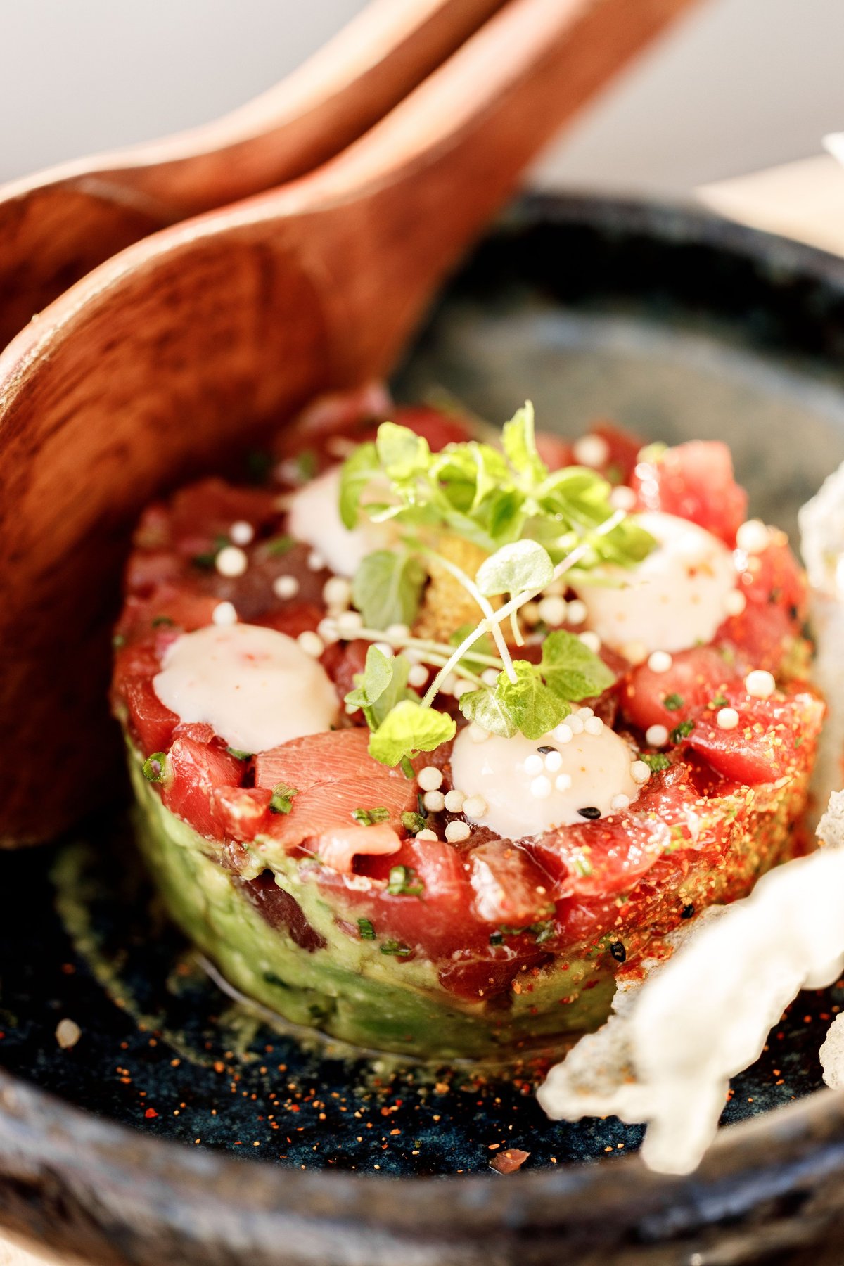 Gourmet tuna tartare appetizer with avocado base, microgreens, and white cream sauce on a dark plate with wooden spoon