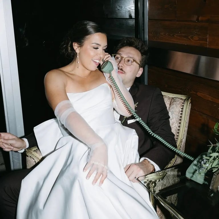Woman in white dress and man in black suit holding a vintage telephone handset together while sitting indoors